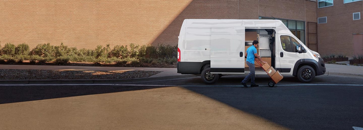 Un hombre empujando paquetes en una carretilla cerca de la puerta lateral corrediza abierta de una Ram ProMaster 3500 SLT Plus Cargo Van Super High Roof 2026 blanca con el paquete de apariencia negro.