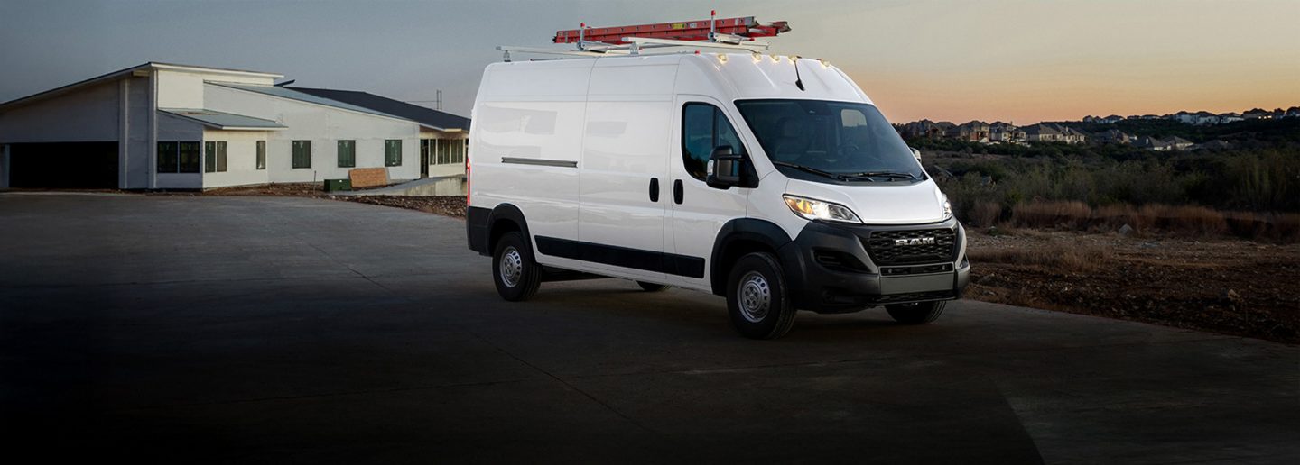 Ángulo frontal del lado del pasajero de una Ram ProMaster Tradesman Cargo Van High Roof 2026 blanca y una escalera en la parrilla del techo saliendo de una obra en construcción.