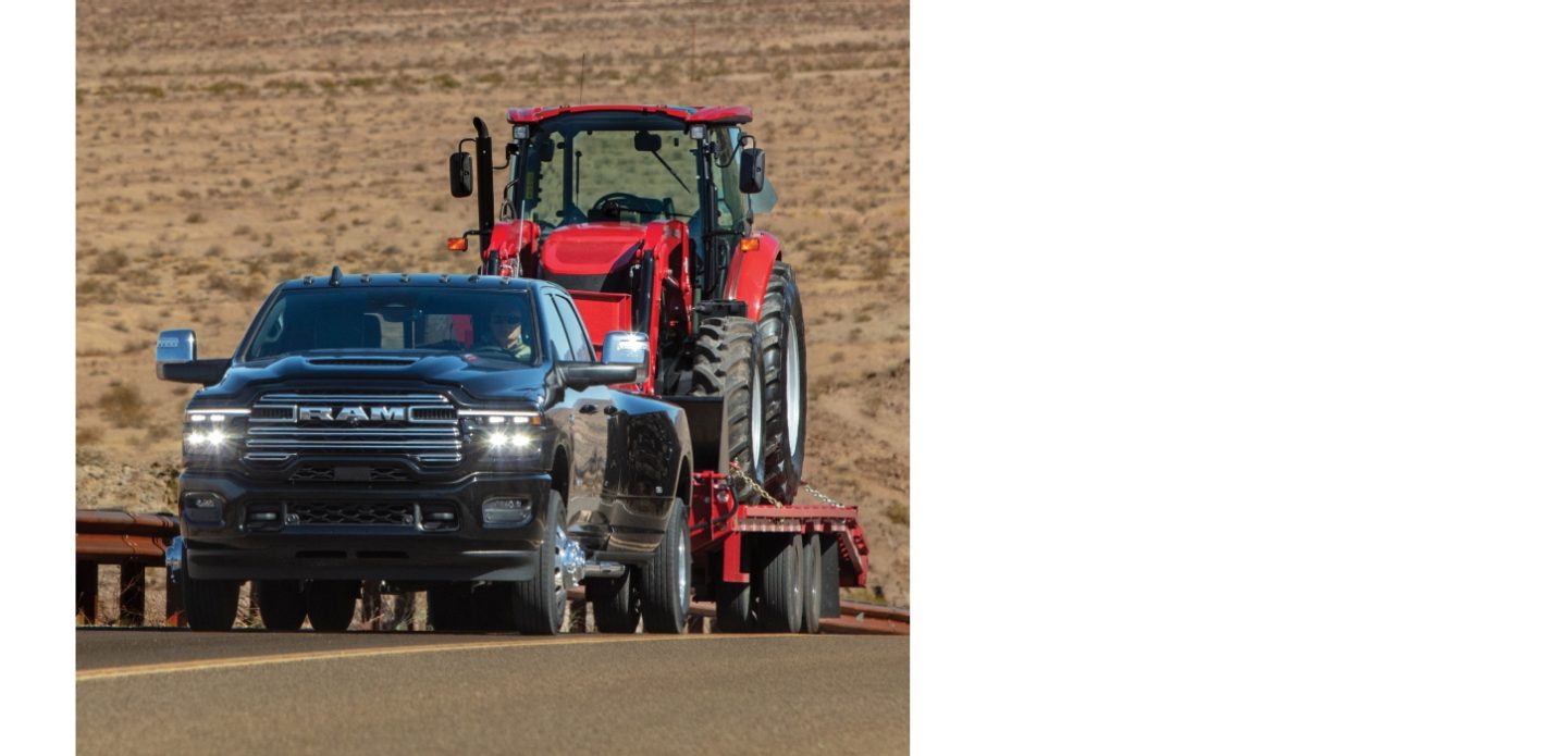 Una pick up de trabajo pesado Ram 3500 Laramie 4x4 Crew Cab 2027 de color azul andando por una autopista en el desierto mientras remolca un tractor Case con un cargador frontal agrícola sobre un remolque de plataforma.