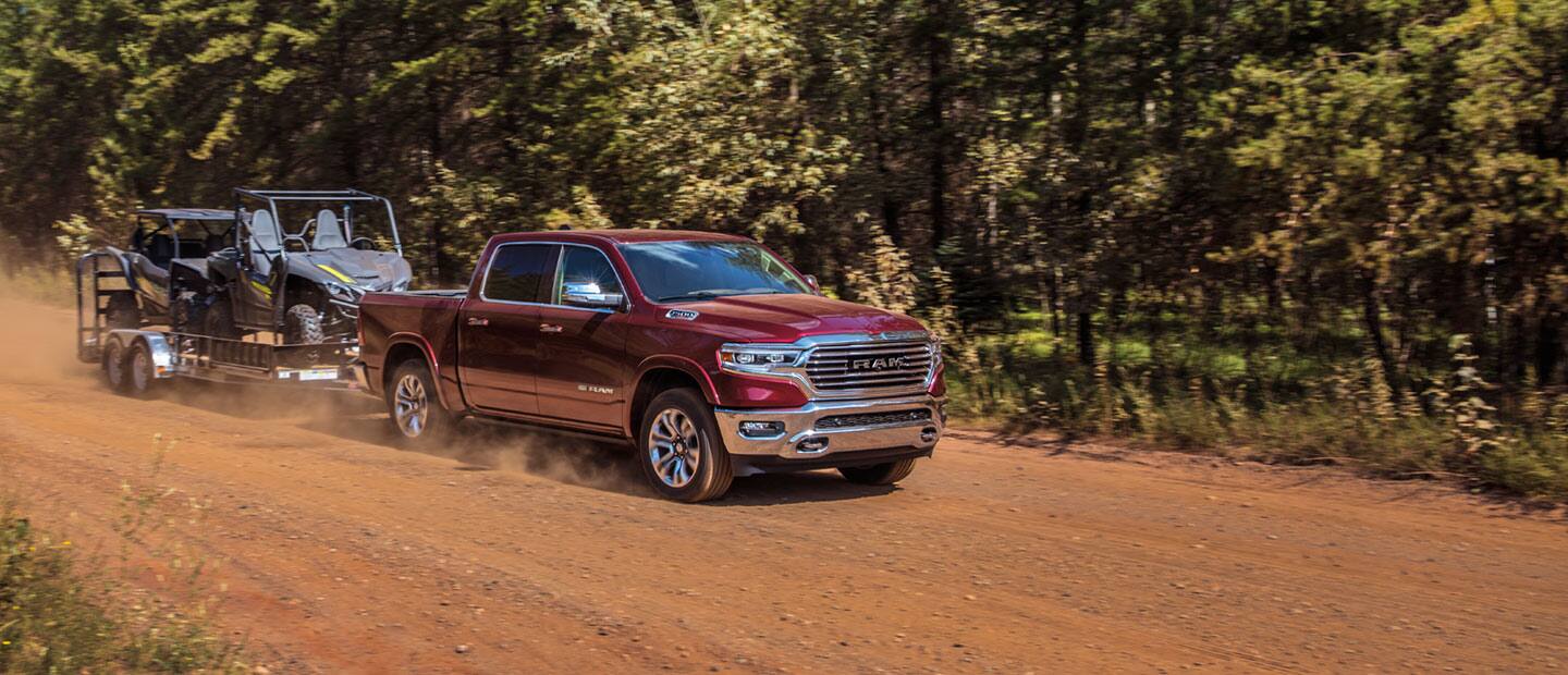 The 2023 Ram 1500 towing two ATVs on a flatbed as it's driven on a dirt road.